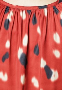 Blusa di seta rossa con pieghe al colletto, decorata con un motivo di grandi gocce nere e crema su uno sfondo uniforme.