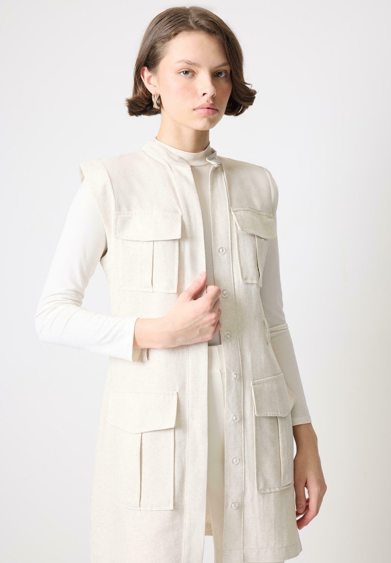 Beige ilma varrukatega vest, mis on valmistatud tekstuuriga kangast, nelja esipocket'iga ja nööbistatud esiosaga. Kantud valge pika varrukaga pluusi peal.