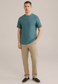 Zöldeskék, textúrázott, rövid ujjú ing vízszintes bordázással, világos bézs nadrággal és sötét cipővel párosítva, semleges háttér előtt áll.