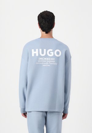 Sweatshirt oversized azul claro com mangas longas. A parte de trás apresenta um grande texto branco "HUGO" e uma marcação adicional por baixo. Tecido suave e texturizado.