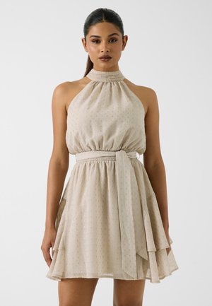 Femme portant une robe beige sans manches à col halter avec de subtils points dorés et une ceinture en tissu assortie, debout devant un fond uni.