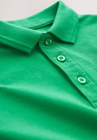 Žalia polo marškinėliai su apykakle, turintys tris sagas ir tekstūruotą audinį. Medžiaga atrodo kvėpuojanti ir lygi.