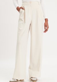 Pantalons à jambes larges de couleur crème avec des plis à l'avant, des poches latérales et une fermeture à un bouton à la taille, associés à des chaussures sportives.