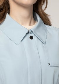 Lichtblauwe jack met een grote kraag, drukknoopsluiting en details van voorzakken. Glad textuur met zichtbare stiksels.
