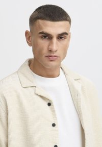 Beige corduroy shirt jacket with a pointed collar and black buttons, layered over a white textured sweater. Short hair. Neutral background.
