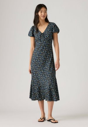 Kvinde iført en navyblå blomster midi-kjole med pufærmer, V-udskæring og sorte flade sandaler, stående på en neutral baggrund.