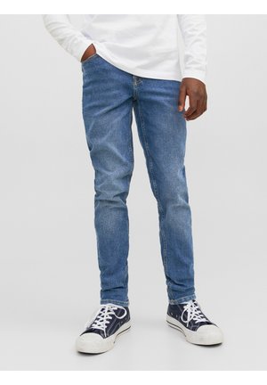 Mand iført blå slim-fit jeans, sorte og hvide sneakers og en hvid langærmet skjorte med den ene hånd i lommen.