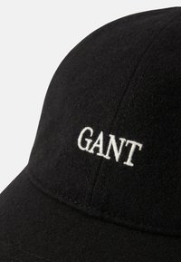 Crna vunena kapa sa strukturiranim oblikom; sadrži bijelu izvezenu oznaku "GANT" sprijeda, suptilnu teksturu i vidljive detalje šivanja.
