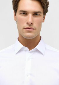 Junger Mann mit heller Haut und braunen Haaren, der ein frisches weißes Hemd trägt, vor einem schlichten hellen Hintergrund.