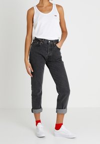 Hvid tanktop med et lille rødt logo, sorte højtaljede jeans med opfoldede nederste kanter, røde sokker og hvide sneakers.