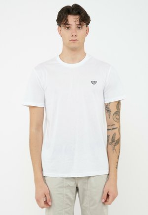 Giovane con capelli ricci che indossa una t-shirt bianca con un piccolo logo nero sul petto e tatuaggi sulle braccia, in piedi davanti a uno sfondo bianco.