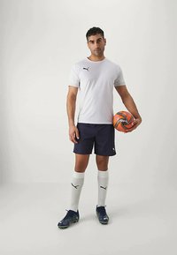 Conjunto deportivo Puma que incluye una camiseta blanca, pantalones cortos nivados, calcetines blancos y botas de fútbol azules. Balón de fútbol naranja y negro en la mano.