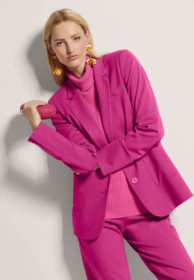 Fuchsia blazer with structured shoulders, paired with a ribbed turtleneck. Accessories include a textured clutch and large gold earrings.