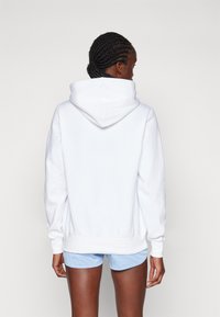 Hvit hettegenser med kengurulomme, teksturert stoff og ribbestrikkede mansjetter. Modellen har på seg lyseblå shorts.