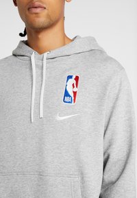 Sweat à capuche gris en matériau doux, avec un swoosh Nike blanc et un logo brodé de la NBA en rouge, blanc et bleu sur la poitrine.