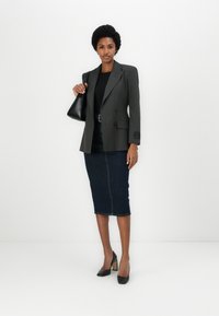 Blazer gris à rayures, double boutonnage, haut noir, jupe crayon en denim foncé, chaussures à talons hauts noires, tenant un sac à main noir.