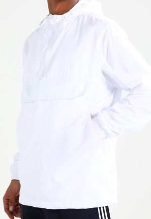 Personne portant un coupe-vent blanc à capuche avec une fermeture éclair sur le devant et les mains dans les poches, associé à un pantalon foncé avec des bandes blanches.