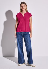 Fuchsiová krátká trička s výstřihem do V a knoflíkovou légou, v kombinaci s volnými modrými denimovými džíny a světlou obuví.