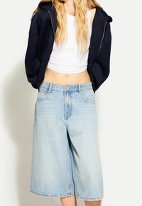 Světlé modré džínové kalhoty culottes s širokými nohavicemi, vysokým pasem a výrazným prošíváním. Kombinované s bílým krátkým tílkem a tmavě modrou mikinou se zipem.