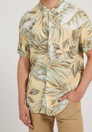 Homme portant une chemise à manches courtes avec une base beige et un motif de feuilles tropicales vertes, associée à un pantalon marron clair.