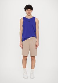 Blå ermeløs tanktop med svart logo, beige shorts med snøring, hvite sokker og hvite joggesko. Enkelt design og avslappet passform.
