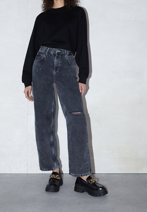 Osoba oblečená v černém topu s dlouhým rukávem, vybledlých černých džínech s vysokým pasem a trhlinou na koleni a robustních černých mokasínách se zlatým řetízkovým detailem.