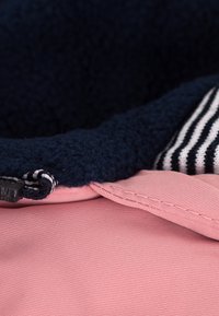 Tessuto esterno rosa con una trama liscia, rivestimento in pile blu navy con una texture morbida e dettaglio a strisce sul bordo. Presenta hardware nero.