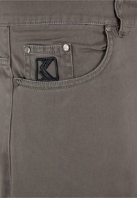 Graue Jeans aus Denim mit strukturiertem Design, einem schwarzen gestickten Logo auf der Gesäßtasche und metallischen Nieten an den Taschenrändern.