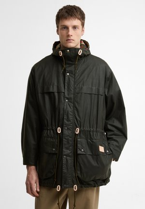 Noor mees, kes kannab tumerohelist kapuutsi ja ees taskutega ning paeladega parkat, seisab ühevärvilise valge tausta ees.