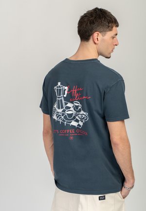 Hombre con camiseta azul oscuro con gráfico y texto temáticos de café "Coffee culture" y "It's coffee o'clock" en la parte trasera, pantalones beige.