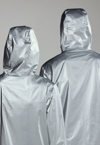 Dos personas con chaquetas plateadas brillantes con capucha, vistas desde atrás sobre un fondo gris liso.