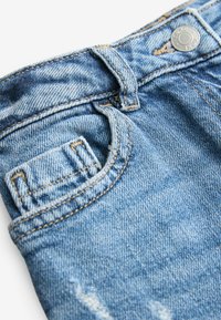 Detailní záběr na světle modré džíny z denimu s přední kapsou, poutkem na opasek a kovovým knoflíkem se švy.