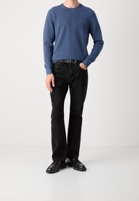 Maglione blu a girocollo, jeans neri a gamba dritta, cintura nera e scarpe nere. Il maglione ha un piccolo logo sul petto sinistro.