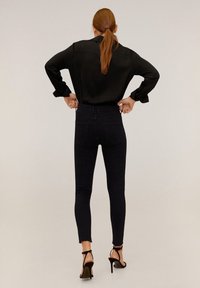 Mujer con cabello rojo, vistiendo una blusa negra y jeans ajustados de cintura alta en negro, con sandalias de tacón alto negras, de pie con las manos en las caderas.