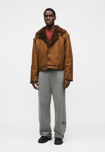Καφέ σουέντ μπουφάν με γούνινο γιακά, με κουμπιά στο μπροστινό μέρος και λεπτομέρειες με ακατέργαστες άκρες, συνδυασμένο με γκρι φόρμα και μαύρα παπούτσια.