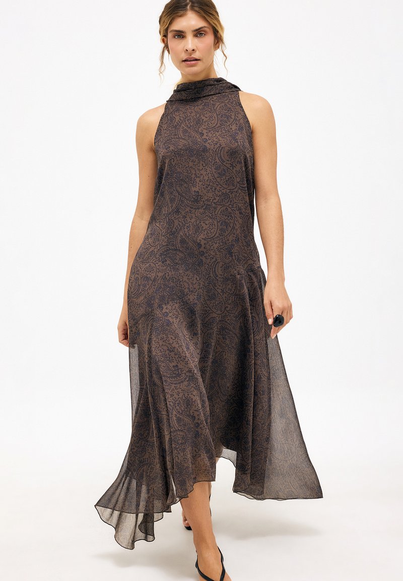 Femme portant une robe midi brune à motifs sans manches, à col haut et ourlet asymétrique, marchant vers l'avant sur un fond uni.