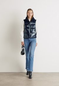 Gilet imbottito blu navy con chiusura a zip, colletto alto e texture lucida, indossato sopra una camicia bianca, abbinato a jeans chiari a zampa d'elefante e stivali neri.