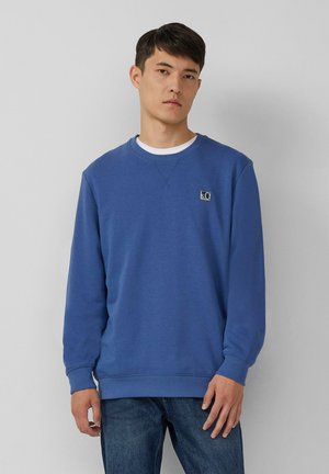 Giovane uomo che indossa un felpa blu con un piccolo logo sul petto e jeans blu, in piedi contro uno sfondo grigio chiaro uniforme.