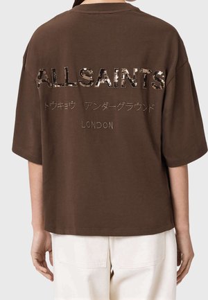 T-shirt marrone oversize con "ALLSAINTS," caratteri giapponesi e "LONDON" stampati sul retro, indossata con pantaloni chiari.