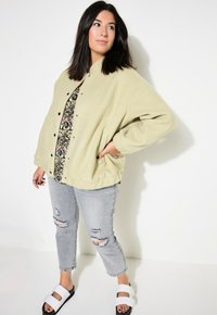 Lichtgroene fleece jacket met zwarte drukknopen, gecombineerd met een patroonshirt, versleten grijze jeans en zwarte slidesandalen.