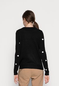 Pull en maille noire avec des accents de points blancs sur les manches, un col rond et des ourlets côtelés. Texture douce avec une coupe décontractée.