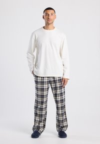 Witte langelmouwen shirt gecombineerd met geruite pyjamabroek in marineblauw, crème en beige. Zachte stof, ontspannende pasvorm en blauwe slippers maken de look compleet.