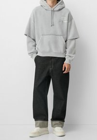 Felpa oversize grigia con tasca frontale e cappuccio con cordino, abbinata a jeans neri taperati con risvolti e sneakers bianche.