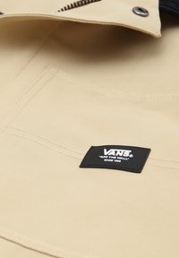 Beige szövet, látható varrással és egy fekete Vans logó címkével varrva. A felület sima és matt textúrájú.