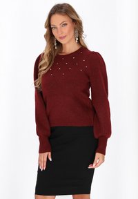 Roter Strickpullover mit Puffärmeln und dekorativen Strasssteinen am Ausschnitt, kombiniert mit einem engen schwarzen Rock. Glatte Textur.