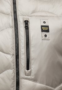 Κοντινό πλάνο υφάσματος ανοιχτόγριου μπουφάν με κάθετο μαύρο φερμουάρ, μία μικρότερη τσέπη με φερμουάρ και μαύρη ετικέτα Blauer U.S.A. με δύο μεταλλικά χοντρά κουμπώματα.