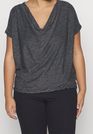 Osoba nosząca czarną i białą bluzkę w paski z krótkimi rękawami oraz czarne spodnie, stojąca na jednolitym tle.