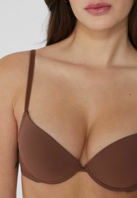 Close-up van een vrouw die een bruine gevoerde BH draagt, waarbij de halslijn, schouder en bovenborst zichtbaar zijn tegen een effen achtergrond.