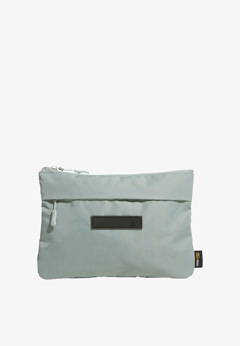 Petite pochette en tissu vert clair avec fermeture à glissière, surface texturée, écusson logo et étiquette latérale. Forme rectangulaire compacte conçue pour le rangement.