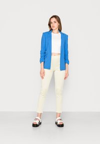 Blazer azul, top branco curto, calças amarelas à quadros e sandálias de plataforma pretas com tiras brancas. Tecido suave e ajuste bem definido.